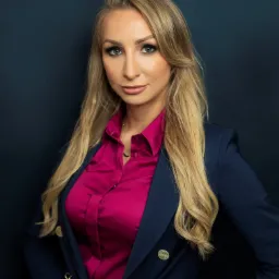 Portret blondwłosej kobiety w granatowej marynarce i fuksjowej koszuli, z delikatnym makijażem i czerwoną biżuterią, pozującej na ciemnym tle.