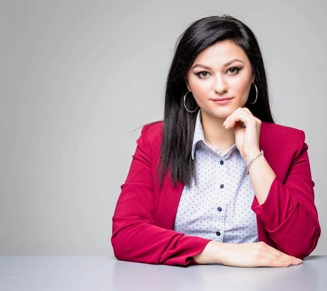 Portret kobiety z ciemnymi włosami w różowym żakiecie, z delikatnym makijażem, opierającej podbródek na dłoni, z kolczykami kołami i bransoletką na nadgarstku.