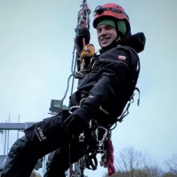 Uśmiechnięty strażak w czerwonym kasku, zawieszony na linach podczas ćwiczeń wysokościowych, z widocznym napisem 'STRAŻ' na kombinezonie i flagą Polski na ramieniu kurtki.