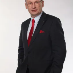 Portret mężczyzny w okularach, ciemnym garniturze w prążki, z czerwoną poszetką i krawatem, na białym tle.