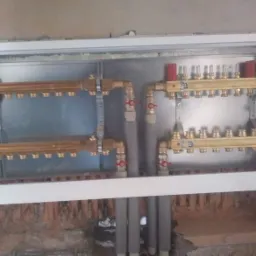 Rozdzielacz centralnego ogrzewania w metalowej szafce, widoczne rury doprowadzające i odprowadzające wodę, oraz zawory regulacyjne.