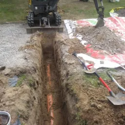 Wykop pod pomarańczowe rury kanalizacyjne, wykonany koparką Volvo, widoczne łopaty i folia ochronna na trawie.