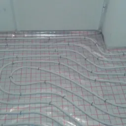 Białe rury ogrzewania podłogowego przymocowane do folii z siatką w pomieszczeniu w trakcie instalacji, widoczne fragmenty ścian.
