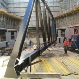 Czarna, stalowa kratownica podnoszona dźwigiem na placu budowy, widoczni pracownicy w kaskach i rusztowania w tle.