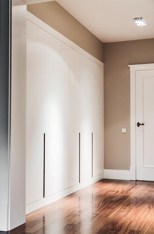 Biała, zabudowana szafa wnękowa z minimalistycznymi, pionowymi uchwytami na tle drewnianej podłogi i beżowej ściany z białymi drzwiami.