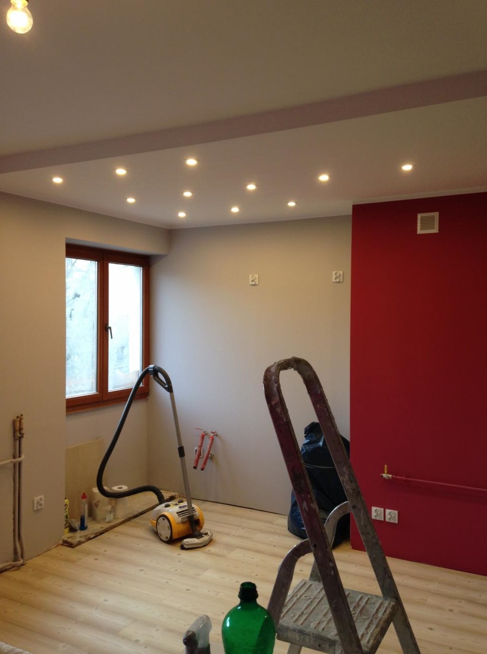 Wnętrze pokoju we Wrocławiu z zamontowanym oświetleniem punktowym w suficie podwieszanym, widoczna drabina, odkurzacz i czerwona ściana w tle.