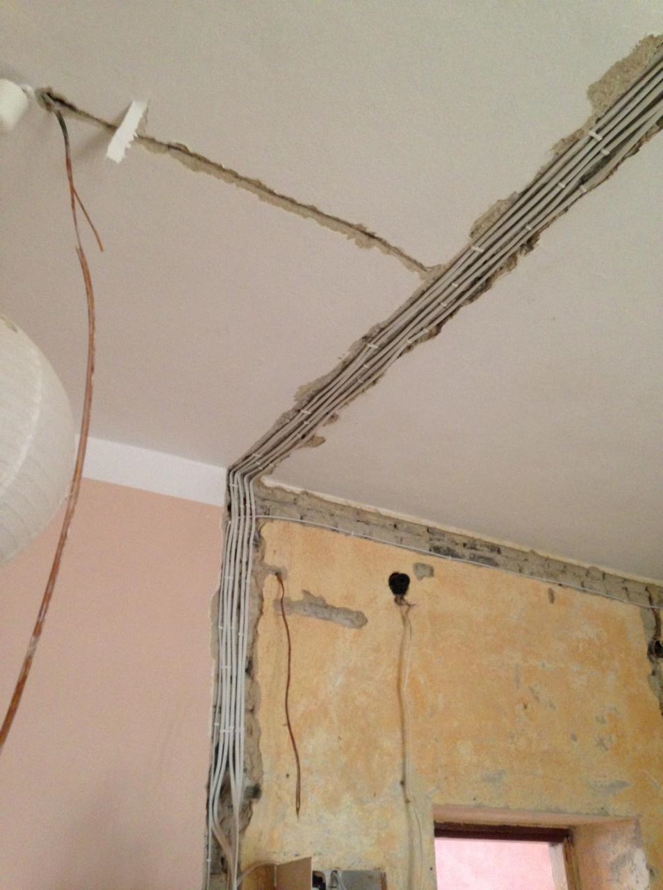 Montaż instalacji elektrycznej: wiązka przewodów w białych peszlach ułożona w wyżłobieniach w suficie i ścianie, mocowana uchwytami, surowy stan wykończenia wnętrza.