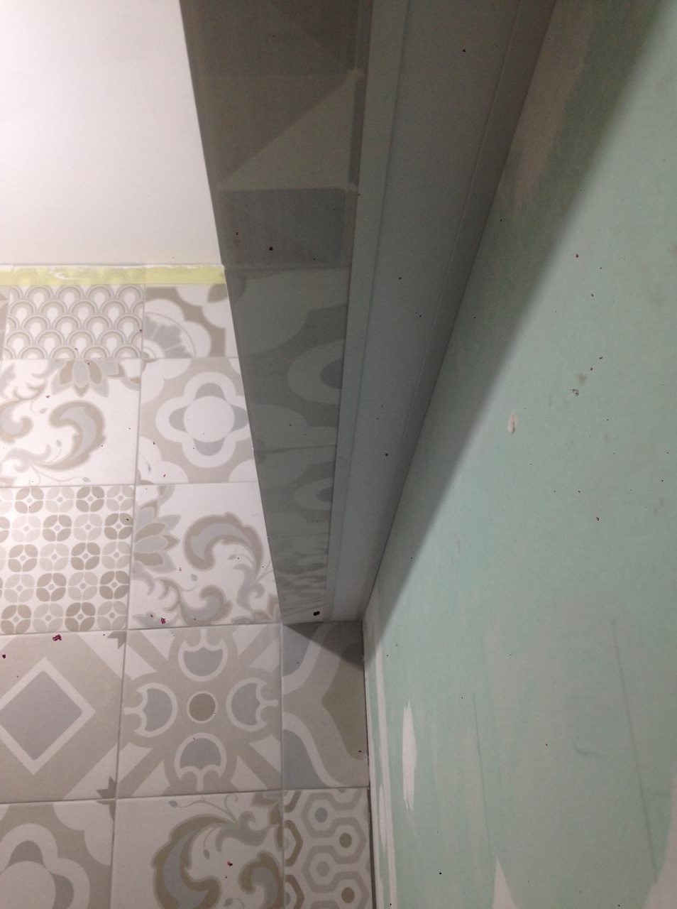 Fragment łazienki w trakcie remontu: dekoracyjne płytki podłogowe, surowa ściana z widocznymi śladami szpachlowania i zamontowana listwa przypodłogowa.