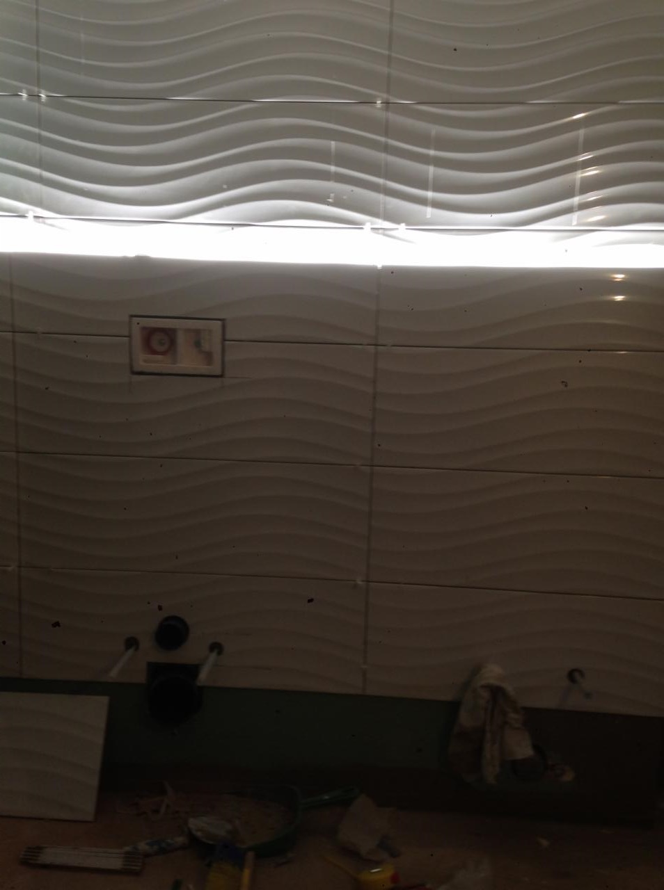 Ściana łazienki z białymi, falowanymi płytkami, widoczne otwory instalacyjne i fragment instalacji podtynkowych oraz narzędzia na podłodze w trakcie prac wykończeniowych.