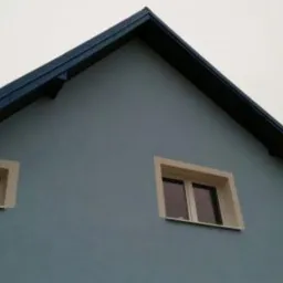 Fragment ściany szczytowej domu jednorodzinnego z dwoma oknami o różnych rozmiarach, pomalowanej na kolor gołębi z ciemnym okapem dachu.