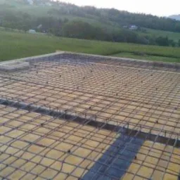 Zbrojenie stropu przygotowane do zalania betonem, widok z góry na siatkę z prętów stalowych, w tle zielone wzgórza i budynek.