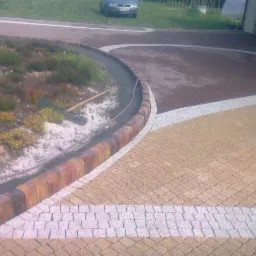 Ułożona nawierzchnia z kostki brukowej w trzech odcieniach, z obrzeżem z cegieł i rabatą kwiatową wzdłuż krawędzi, widok z góry, na rabacie leży grabie.