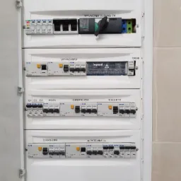 Otwarta skrzynka elektryczna z bezpiecznikami i wyłącznikami oznaczonymi nazwami obwodów, widoczne narzędzie pomiarowe w dolnej części obudowy.