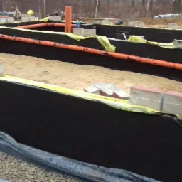 Przygotowany wykop pod fundamenty z izolacją, rurami kanalizacyjnymi i częściowo ułożonymi bloczkami betonowymi.