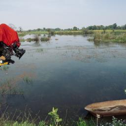 Paweł Kacprzak-produkcja filmowa, realizacja filmów reklamowych, promocyjnych, dokumentalnych - Operator kamery z osłoniętą kamerą filmową na wysięgniku nad wodą z łodzią w tle, podczas kręcenia plenerowej sceny.