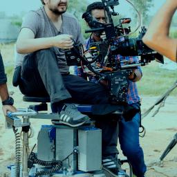 Paweł Kacprzak-produkcja filmowa, realizacja filmów reklamowych, promocyjnych, dokumentalnych - Operator kamery siedzący na wózku kamerowym z dużą kamerą filmową, drugi operator za nim, na planie filmowym z polnym tłem.