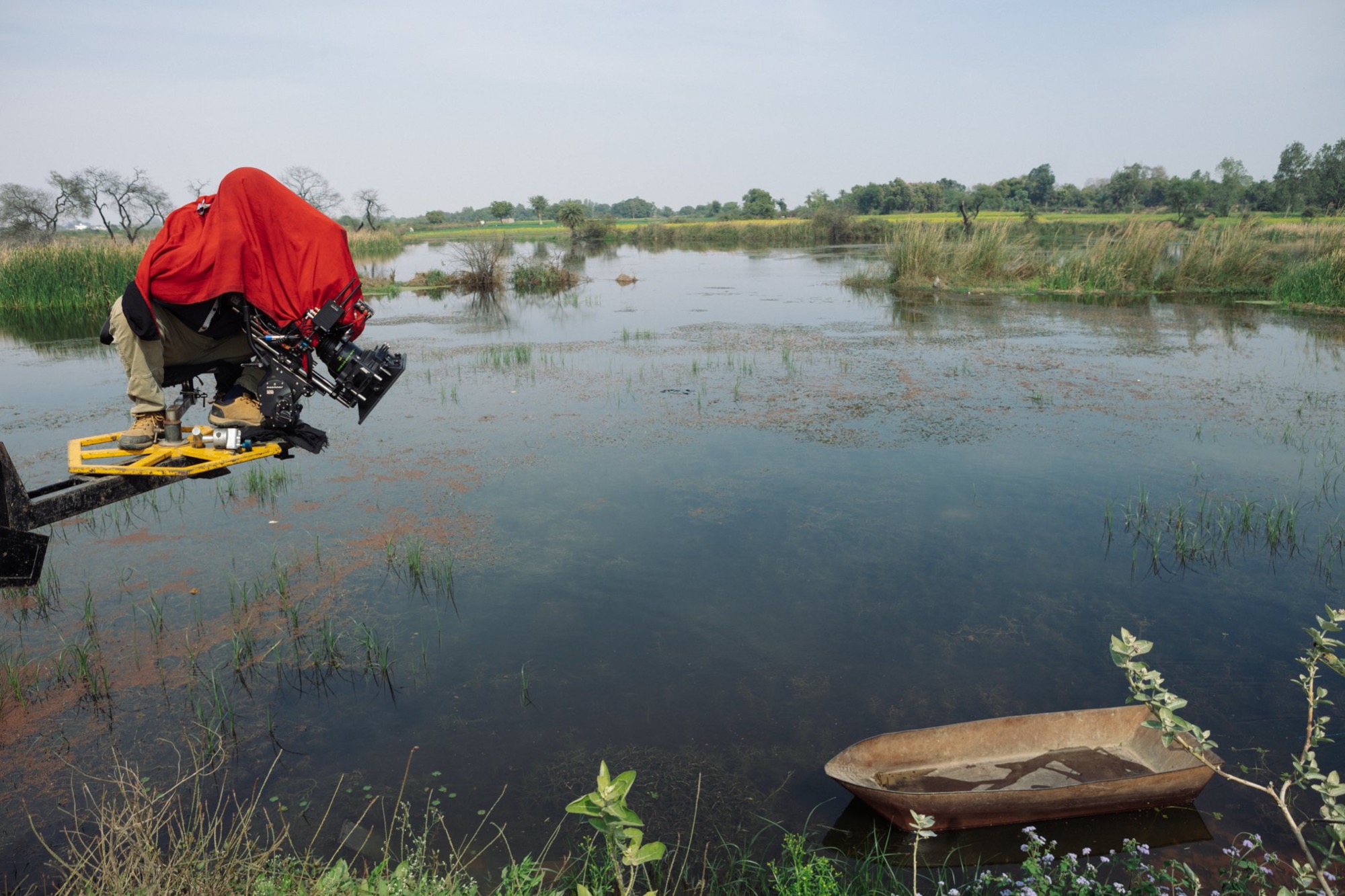 Operator kamery z osłoniętą kamerą filmową na wysięgniku nad wodą z łodzią w tle, podczas kręcenia plenerowej sceny.