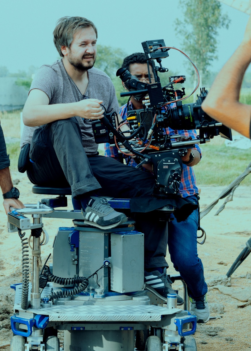 Operator kamery siedzący na wózku kamerowym z dużą kamerą filmową, drugi operator za nim, na planie filmowym z polnym tłem.