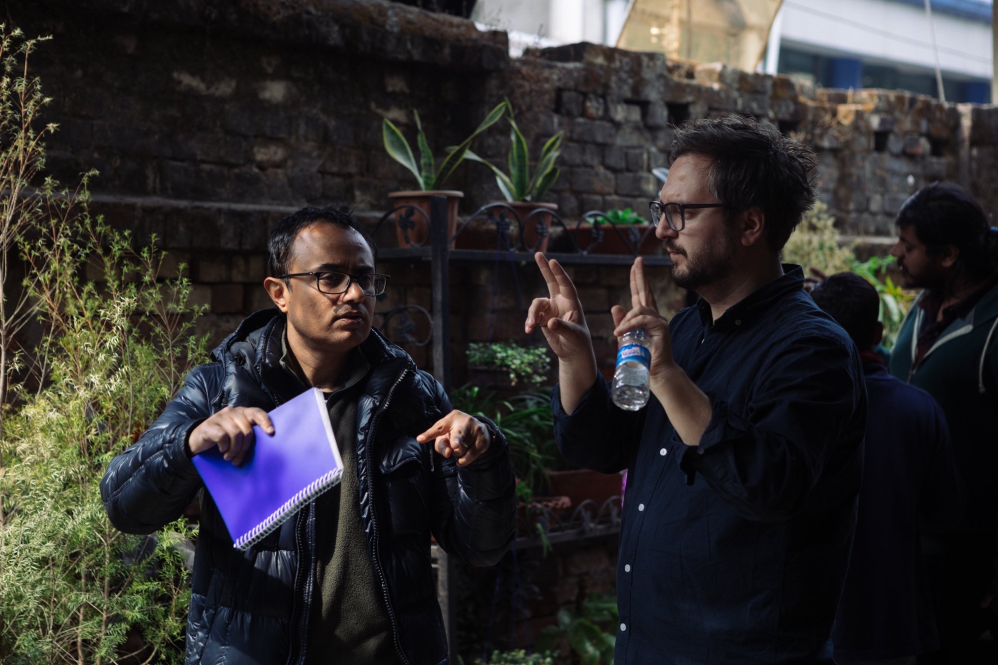 Dwaj mężczyźni w okularach dyskutują o planie filmowym na tle ceglanej ściany z roślinami doniczkowymi; jeden trzyma niebieski notatnik, drugi gestykuluje z butelką wody.