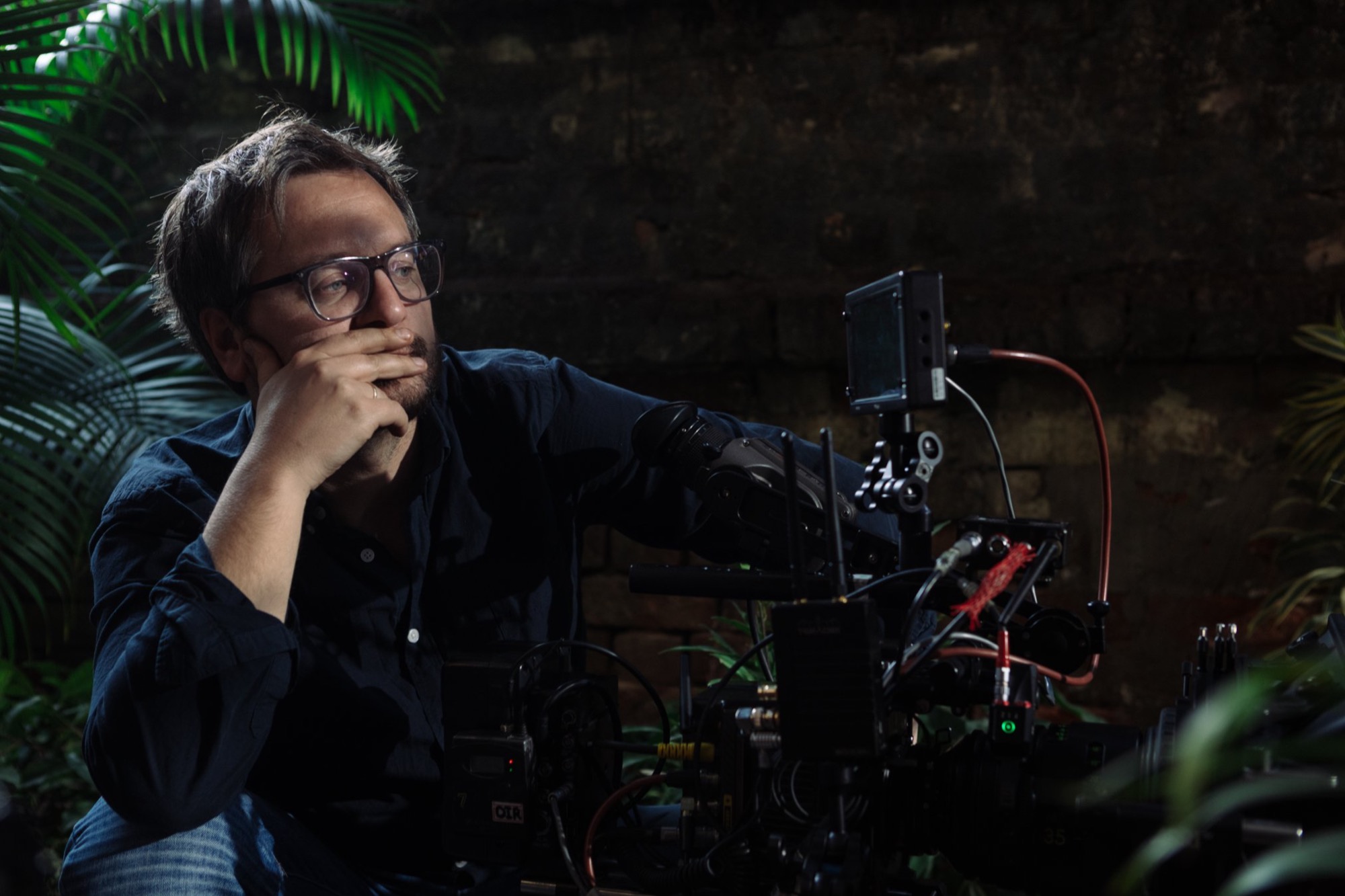 Operator filmowy w okularach, zamyślony, oparty o kamerę filmową w otoczeniu roślin tropikalnych i ściany z cegły.