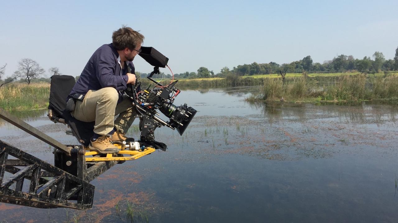 Operator kamery na platformie wysięgnika filmuje bagienny krajobraz z trzcinami i drzewami w tle.