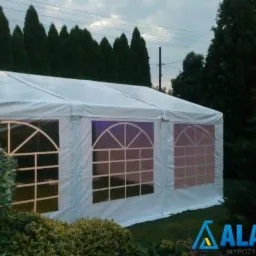 Biały namiot z oknami w ogrodzie, widoczne logo firmy AlanTent w prawym dolnym rogu.