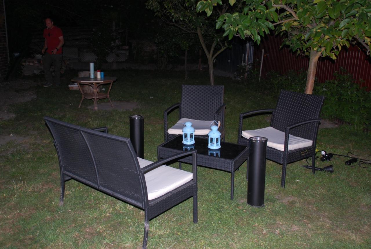 Ciemne, rattanowe meble ogrodowe z kremowymi poduszkami na trawie, oświetlone lampionami i lampkami, z mężczyzną w tle.