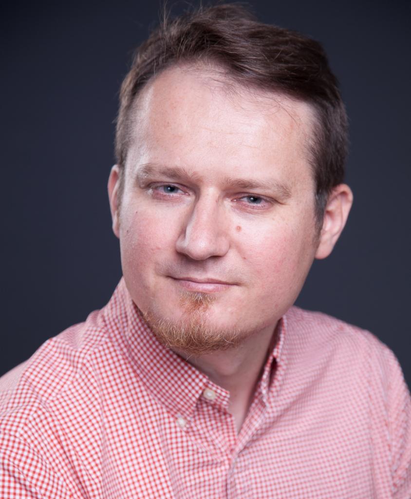 Portret mężczyzny w czerwonej koszuli w kratę z krótką bródką, na ciemnoszarym tle.