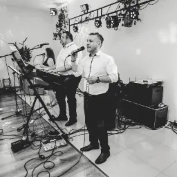 Czarno-białe zdjęcie zespołu muzycznego podczas występu: wokalista z mikrofonem, klawiszowiec grający na keyboardzie, oświetlenie sceniczne i sprzęt nagłośnieniowy.