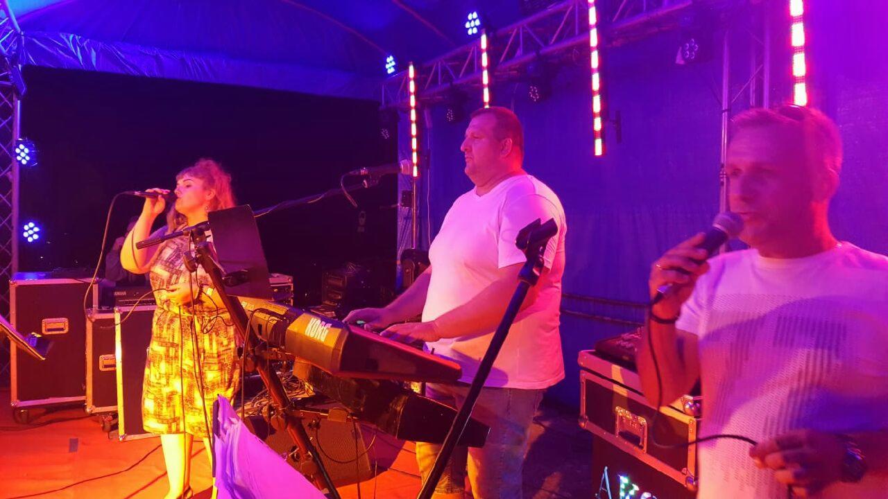 Zespół muzyczny podczas występu na scenie, wokalistka w sukience z mikrofonem, klawiszowiec grający na Korgu, wokalista śpiewający do mikrofonu, oświetlenie sceniczne w tle.