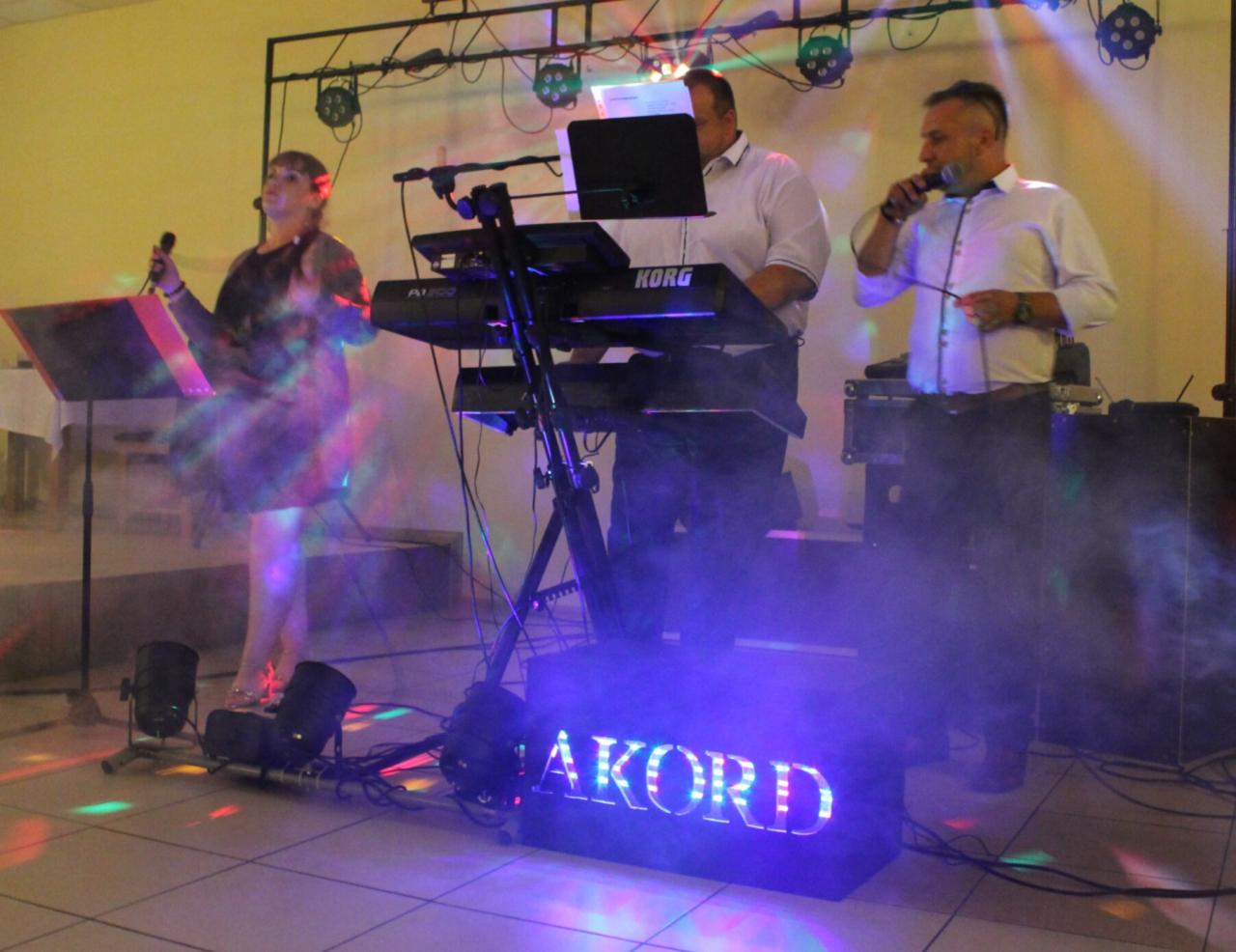 Występujący na scenie muzycy: wokalistka, klawiszowiec grający na Korgu i wokalista z mikrofonem, oświetleni reflektorami i efektem dymu, z podświetlanym napisem 'AKORD' z przodu.