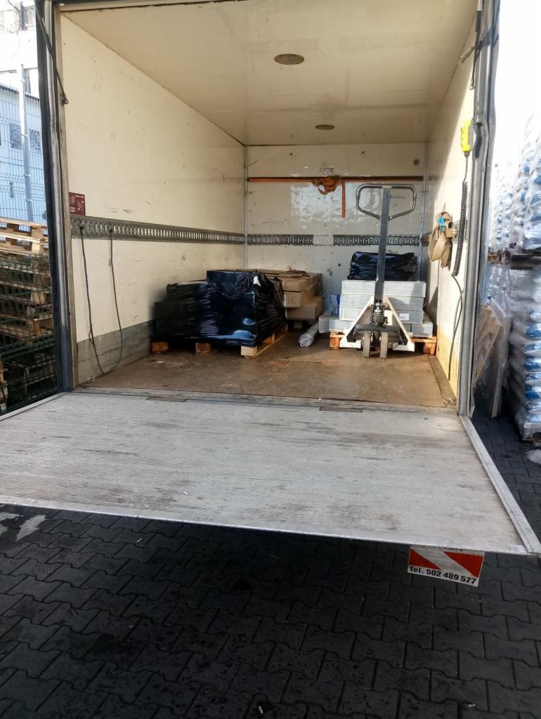 Wnętrze otwartego busa transportowego z podnośnikiem hydraulicznym, paletami z towarem zabezpieczonym folią i wózkiem paletowym w środku.