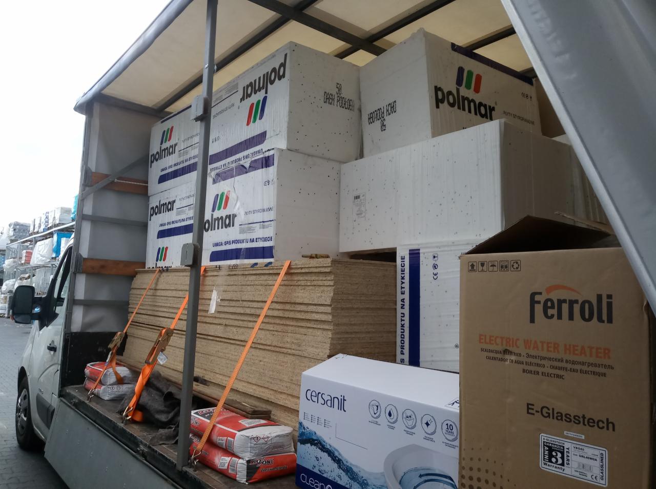 Wnętrze busa transportowego załadowane materiałami budowlanymi: płyty drewniane, styropian Polmar, opakowania Cersanit i Ferroli, zabezpieczone pasami transportowymi.