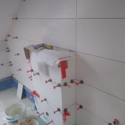 RBM Mariusz Bonk - Świeżo ułożone białe płytki na ścianie z systemem poziomowania płytek i czerwonymi klinami, widoczne również wiaderka i plany.