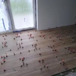 Układanie drewnopodobnych płytek podłogowych z użyciem systemu poziomowania, widoczne krzyżyki dystansowe i kliny w kolorze pomarańczowym, okno zabezpieczone folią malarską.