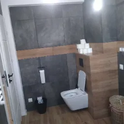 Nowoczesna łazienka z szarymi płytkami imitującymi beton, drewnopodobnymi panelami, wiszącą toaletą i grzejnikiem drabinkowym.