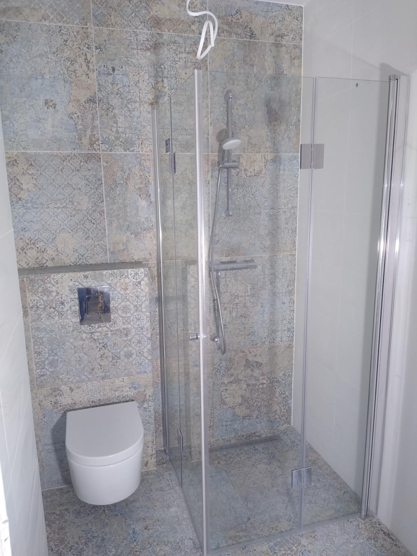 Wykończona łazienka w Koleczkowie: kabina prysznicowa ze szklanymi drzwiami, wisząca toaleta i wzorzyste płytki w odcieniach szarości i beżu.