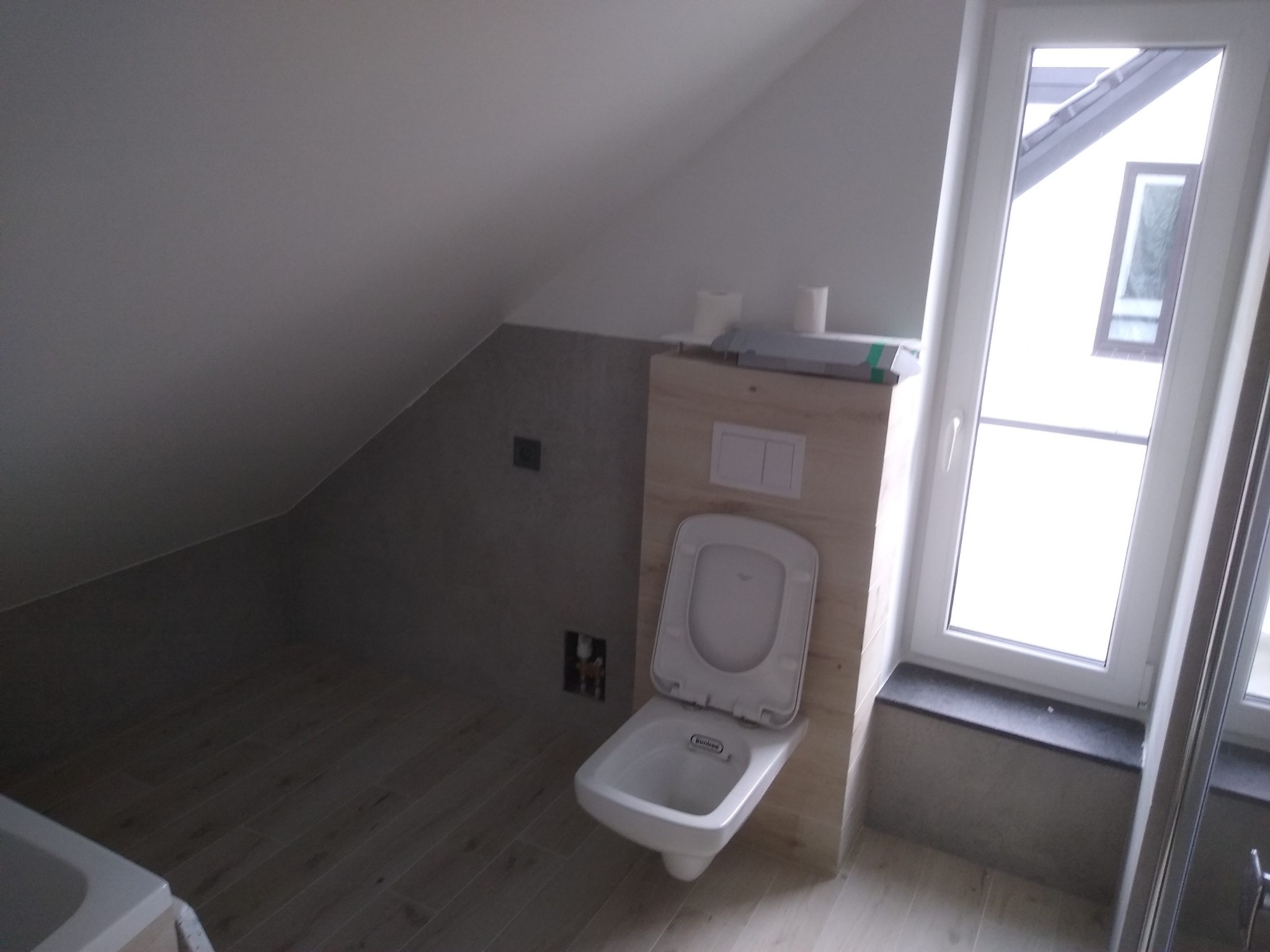 Podwieszana toaleta z deską w jasnej łazience na poddaszu, widok na okno dachowe