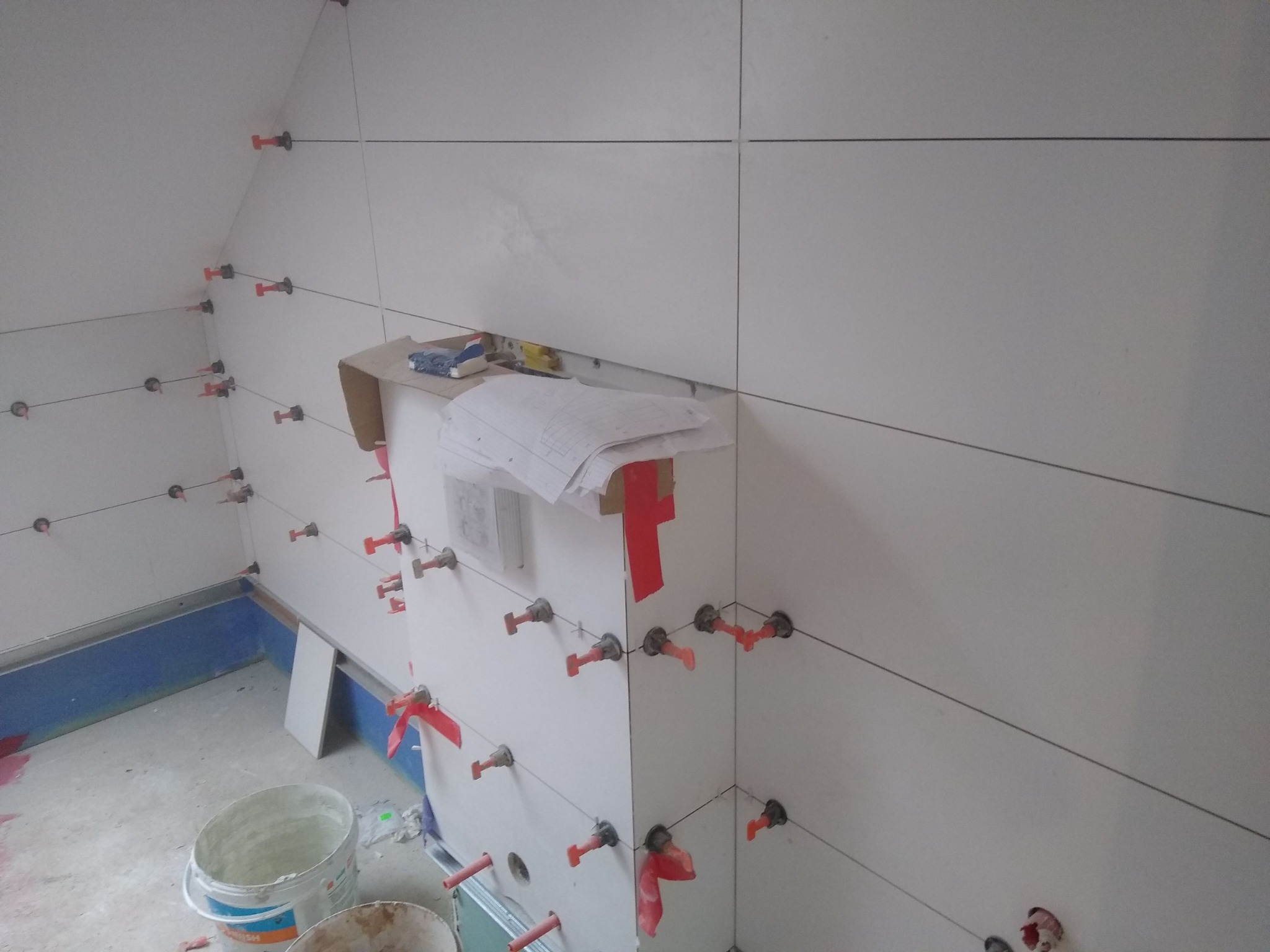 Świeżo ułożone białe płytki na ścianie z systemem poziomowania płytek i czerwonymi klinami, widoczne również wiaderka i plany.