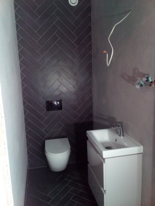 Mała łazienka z ciemnymi, ułożonymi w jodełkę płytkami na ścianie i podłodze, białą umywalką z szafką i wiszącą toaletą, widoczne niedokończone podłączenia elektryczne.