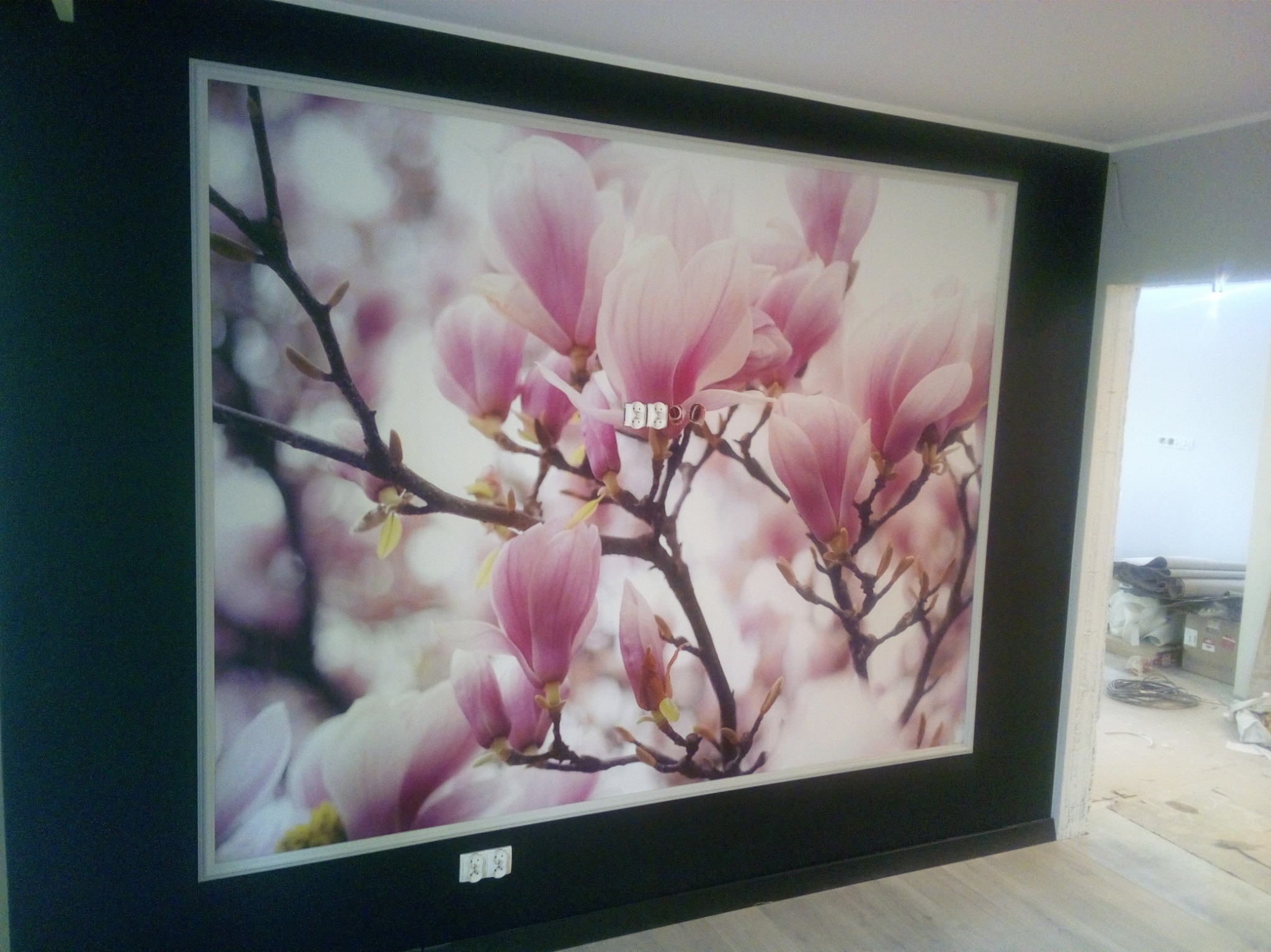 Ściana z fototapetą przedstawiającą kwitnące magnolie w odcieniach różu, otoczona czarną ramą, widok na remontowane pomieszczenie w tle.