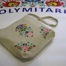 Beżowa torba z pł&oacute;tna ozdobiona trzema haftowanymi bukietami kwiat&oacute;w w stylu ludowym, na białym tle z częściowo widocznym, kolorowym motywem kwiatowym i napisem 'POLYM', widok z g&oacute;ry.