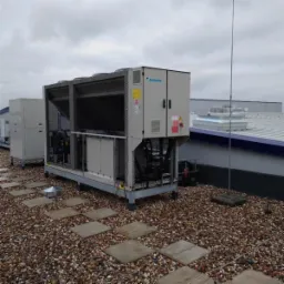 Agregat chłodniczy Daikin na dachu pokrytym żwirem, widoczna instalacja odgromowa i inne urządzenia w tle.