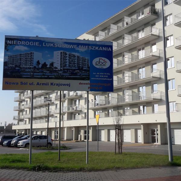 Billboard reklamujący nowe apartamenty w Nowej Soli, widoczny budynek z balkonami i garażami, zaparkowane samochody, trawnik.