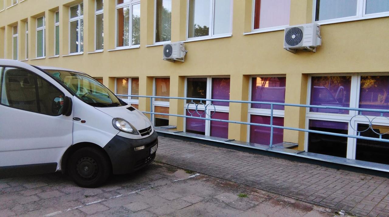 Żółty budynek z zamontowanymi jednostkami zewnętrznymi klimatyzacji, biały samochód dostawczy zaparkowany przed budynkiem, widoczne fioletowe folie na oknach.