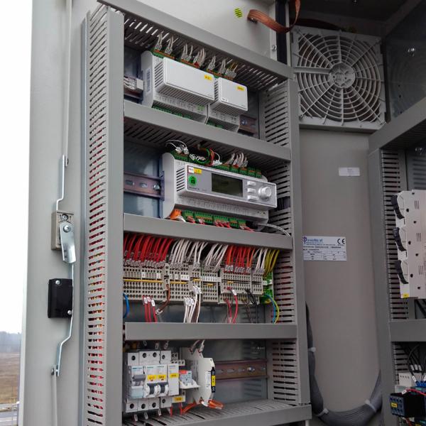 Otwarta szafa sterownicza z licznymi przewodami, bezpiecznikami, wentylatorem i modułami elektronicznymi, widok z boku na szarą obudowę z zamkiem.