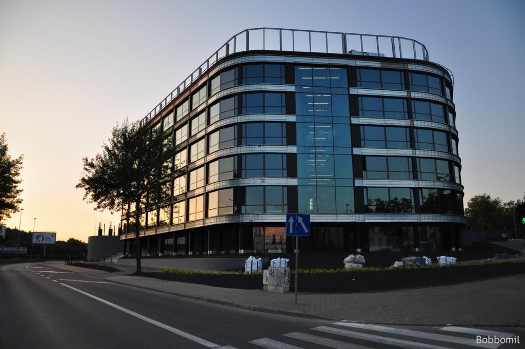 Nowoczesny, przeszklony budynek biurowy z widocznymi instalacjami wentylacyjnymi na dachu, sfotografowany o zmierzchu z ulicy.