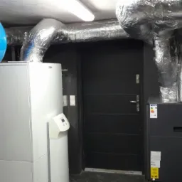 Wnętrze kotłowni z zainstalowanym systemem urządzeń: biały zasobnik, niebieski zbiornik ciśnieniowy, szare urządzenie oraz srebrne rury izolowane termicznie pod sufitem.