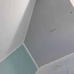 Świeżo pomalowane wnętrze z widocznymi różnymi odcieniami szarości i błękitu na ścianach i suficie, puszki z farbą na podłodze, widoczne gniazdka elektryczne.