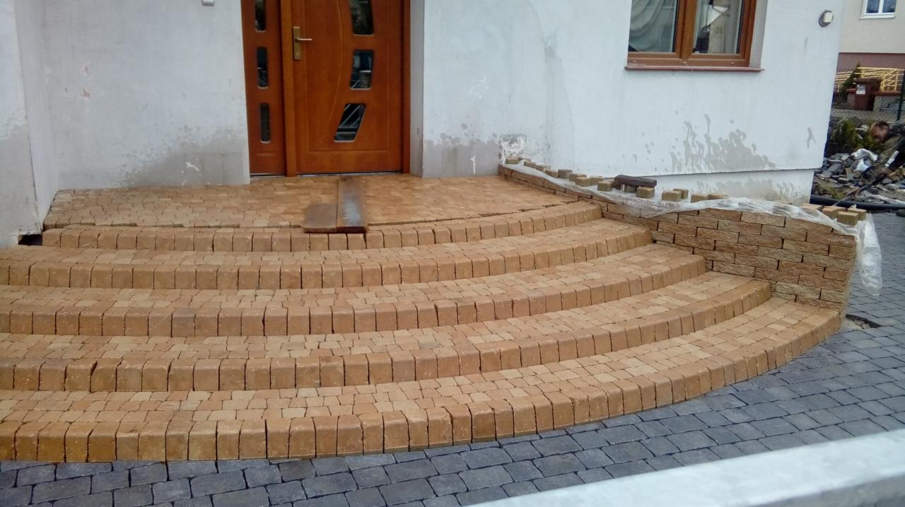 Schody wejściowe do domu w trakcie układania z kostki brukowej w odcieniach beżu i brązu, widoczny fragment podjazdu z szarej kostki, prace w toku.
